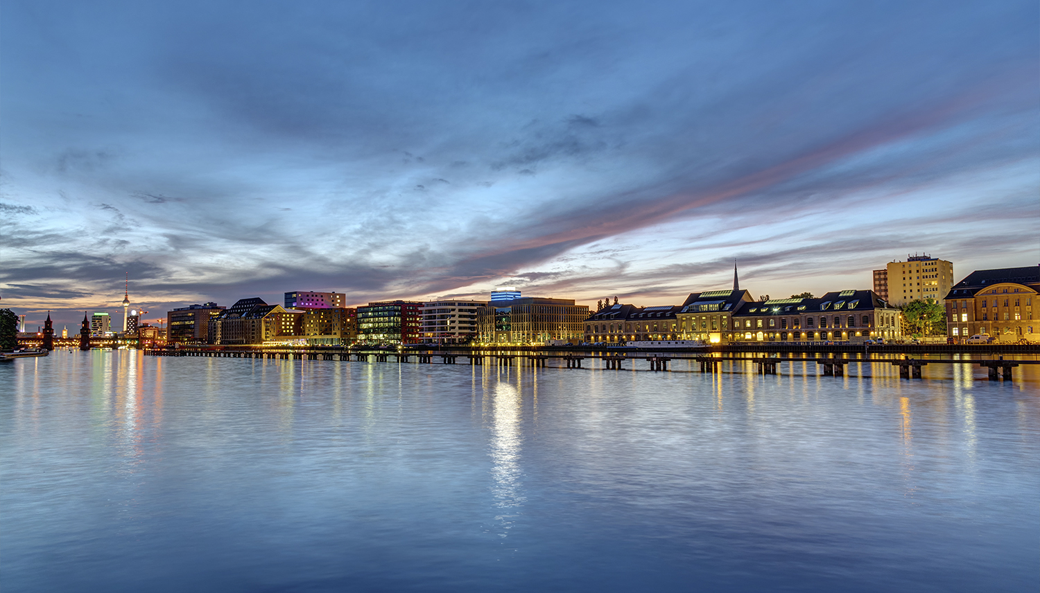 Upplysta byggnader längs floden Spree i Berlin i skymningen.