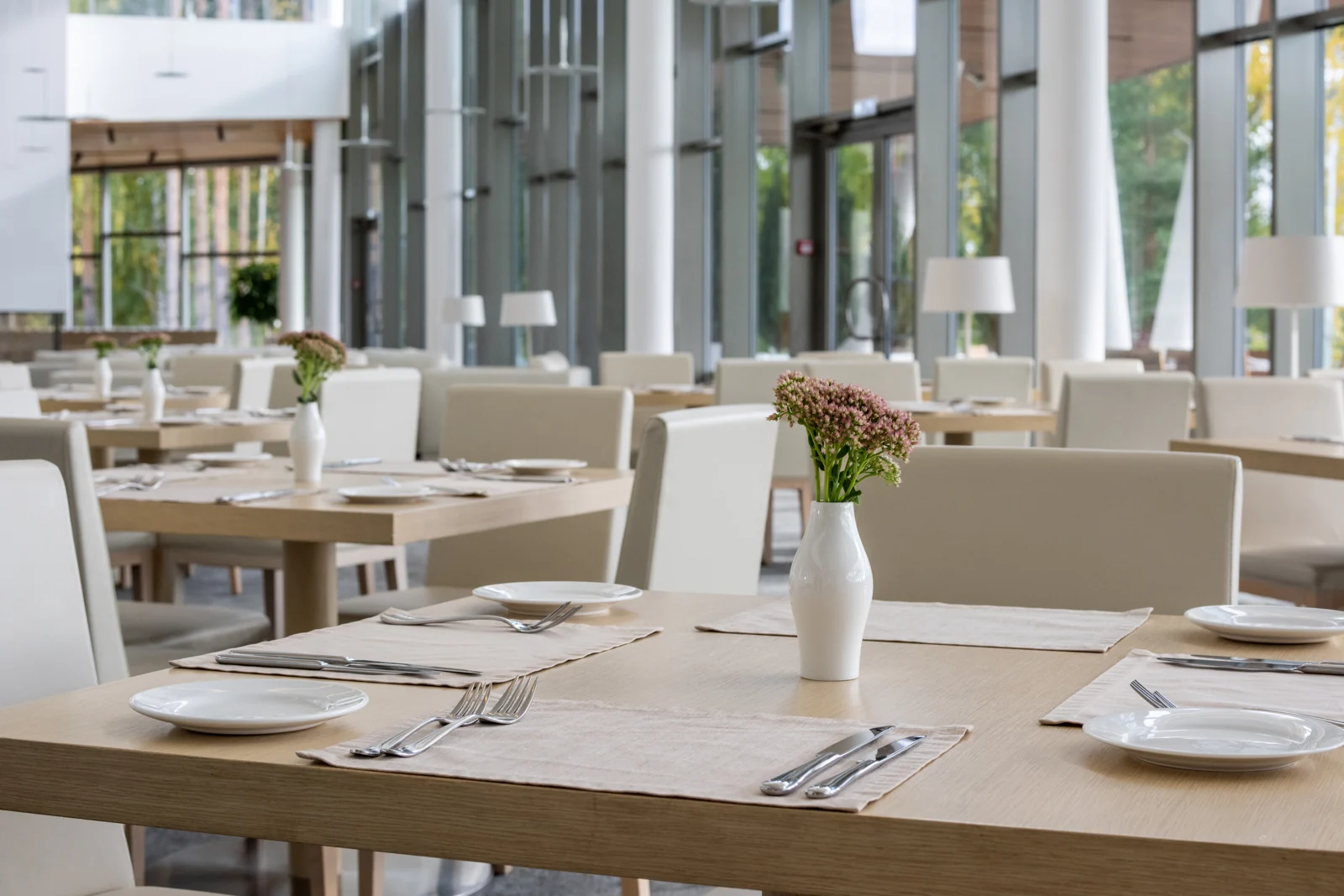 Dukade bord med ljusa stolar och små blomvaser i en restaurang med stora fönster.